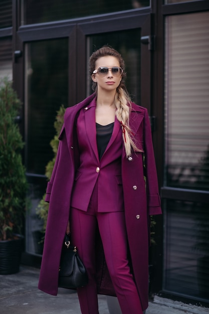 Foto de archivo retrato de mujer de negocios elegante con trenza en gafas de sol, vistiendo un traje morado brillante de moda y una gabardina sobre los hombros. Ella sostiene un lujoso bolso de cuero en la mano.