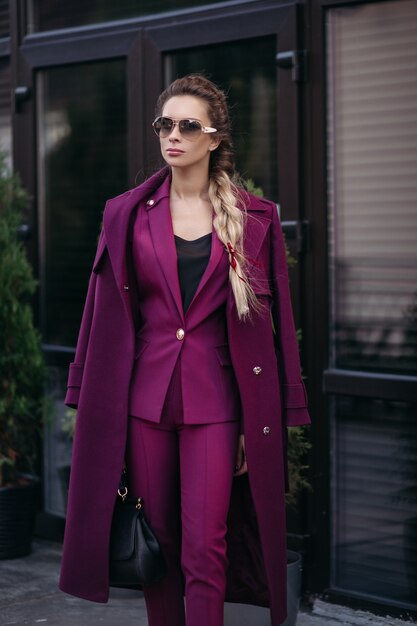 Foto de archivo retrato de mujer de negocios elegante con trenza en gafas de sol, vistiendo un traje morado brillante de moda y una gabardina sobre los hombros. Ella sostiene un lujoso bolso de cuero en la mano.