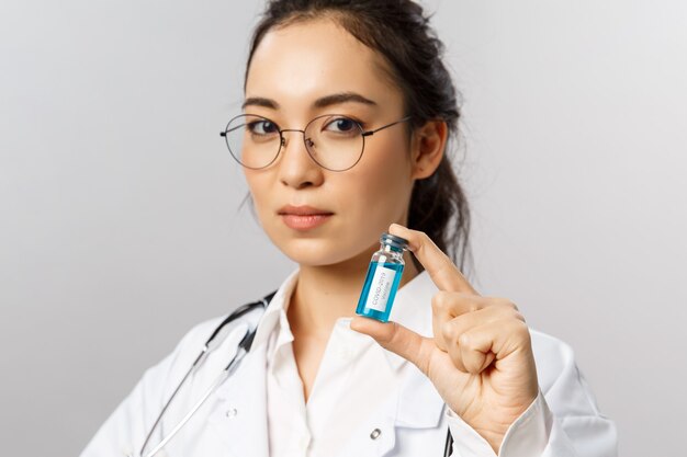 Foto de archivo retrato de primer plano de una joven doctora asiática de aspecto serio que muestra una ampolla con corona ...