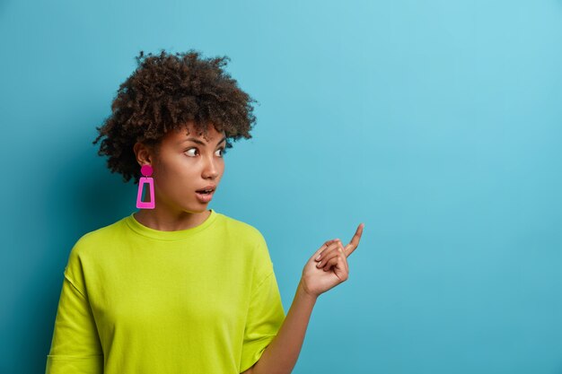 La foto de la asombrada mujer de pelo rizado apunta al espacio de la copia lateral con expresión de asombro, muestra una pared en blanco para la presentación de la idea, vestida de manera informal demuestra una locura