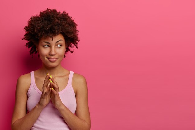 Foto de astuta adolescente rizada que empina los dedos, trama algo, inventa una buena idea, reflexiona sobre algo, mira misteriosamente a un lado, usa una camisa informal, posa en interiores sobre una pared rosa