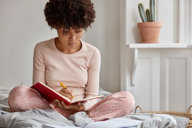 La foto de una atractiva dama negra en ropa casual escribe una historia en un cuaderno con un bolígrafo, mantiene las piernas cruzadas, posa en la cama, hace la planificación para la próxima semana, modelos en el dormitorio. Imagen suave. Estilo de vida