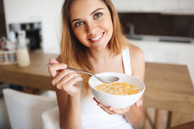 Foto de atractiva jovencita comiendo copos de maíz con leche en la cocina sonriendo