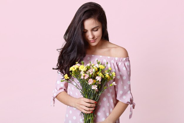 La foto de la atractiva modelo femenina morena tiene flores de primavera, usa un vestido de lunares