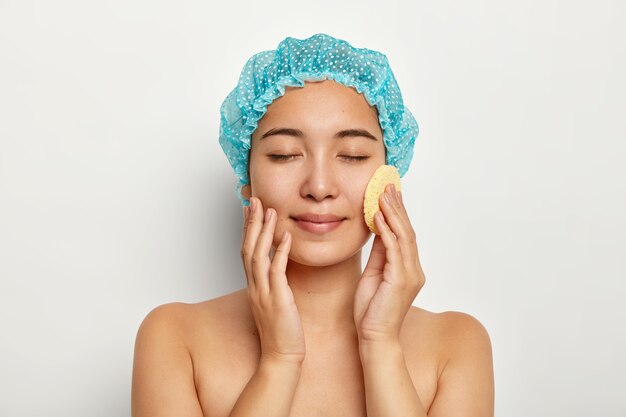Foto de atractiva mujer asiática que se lava la cara con una esponja cosmética, limpia la cara, se para en topless, mantiene los ojos cerrados, viste un gorro de baño azul