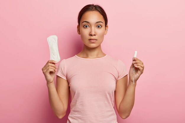 La foto de una atractiva mujer asiática sostiene un tampón de algodón y una toalla sanitaria, tiene una mirada de sorpresa, tiene un ciclo menstrual mensual y usa una camiseta rosa. Cuidado protector para la salud de la mujer. Concepto de fertilización