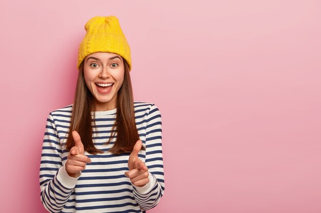 La foto de la atractiva mujer joven moodel hace un gesto de pistola con los dedos, viste un sombrero amarillo y un jersey a rayas, expresa su elección, te elige para su equipo, aislado sobre fondo rosa.