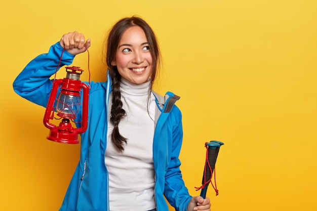 Foto de una atractiva mujer morena con expresión pensativa y alegre, sostiene una lámpara de gas roja, bastones de trekking, cubre largas distancias en un sendero de montaña, usa cuello alto y chaqueta azul, recuerda un momento agradable