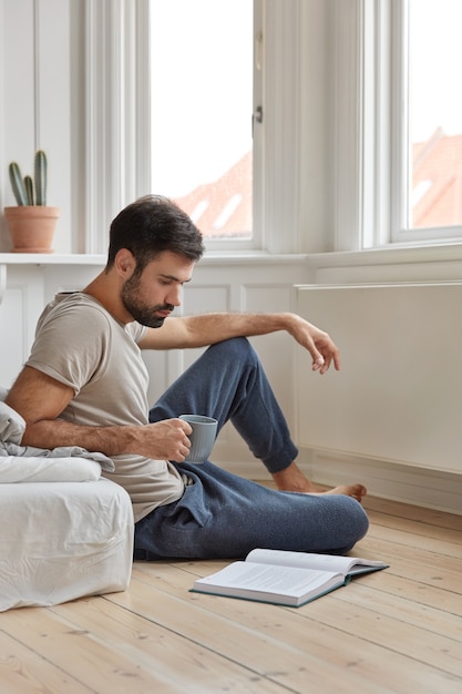 Foto de atractivo hombre inteligente disfruta leyendo un libro en casa, se sienta en el piso cerca de la cama, bebe bebidas calientes frescas, le gusta la novela, se siente inspirado y relajado, disfruta de un ambiente tranquilo. La literatura nos desarrolla