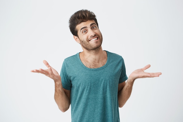 Foto de un atractivo hombre sonriente con barba vestido con una camiseta azul, diciendo que no sabe qué hacer. Estudiante elegante, desconcertado y desconcertado, encogiéndose de hombros expresando incertidumbre. Expreso facial