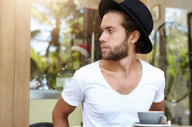 Foto de atractivo joven con barba elegante sentado en el café, mirando a otro lado, tratando de ver al camarero