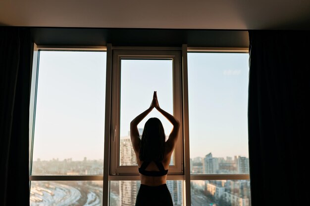 La foto de atrás de una dama delgada y bonita en la parte superior del deporte está haciendo movimientos de yoga frente a la ventana bajo el sol en la mañana en casa