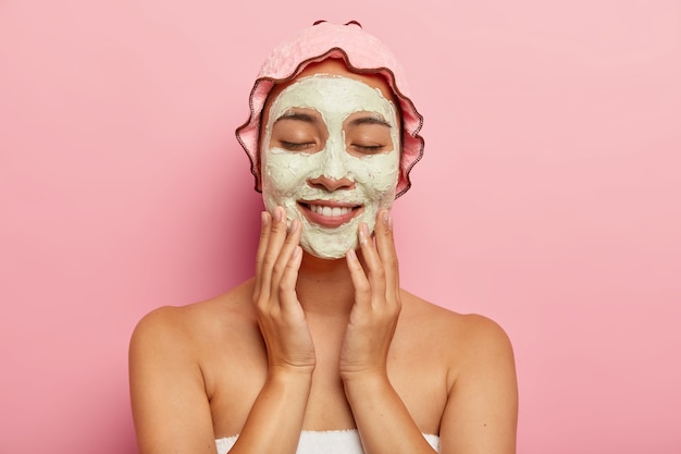 Foto de belleza de sonriente joven étnica aplica mascarilla hidratante en la cara, se somete a un tratamiento facial en el interior, se para con los hombros desnudos, usa un sombrero de ducha rosa en la cabeza