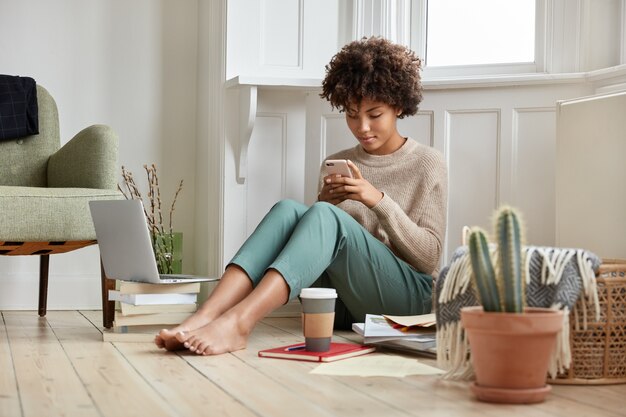 Foto de una bonita niña afro sentada en una acogedora habitación en el piso, busca el perfil en las redes, toma café, trabaja con literatura y computadora portátil, conversa en línea en el teléfono móvil, usa un suéter informal y pantalones