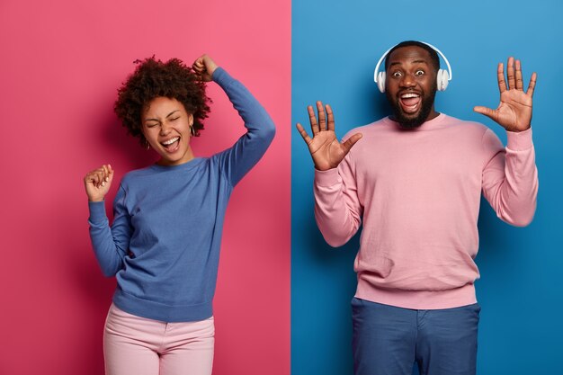 Foto brillante de una pareja afroamericana complacida enérgica baila con los brazos levantados, pasa el tiempo libre en la fiesta disco, usa auriculares