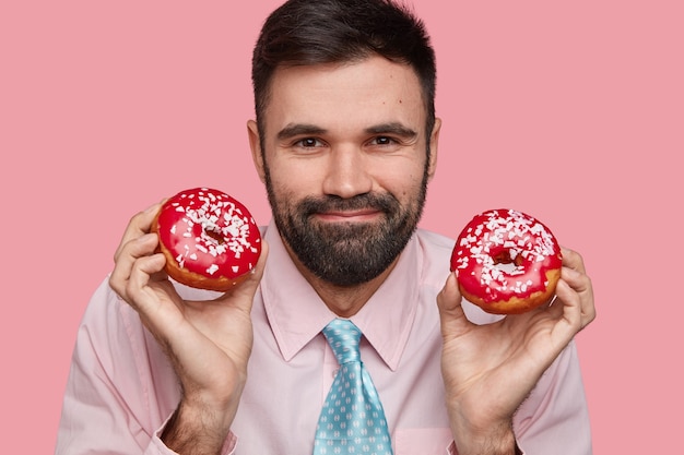 La foto en la cabeza del atractivo hombre barbudo tiene una expresión amistosa, una sonrisa suave, cerdas gruesas, usa camisa y corbata, sostiene deliciosas donas en ambas manos