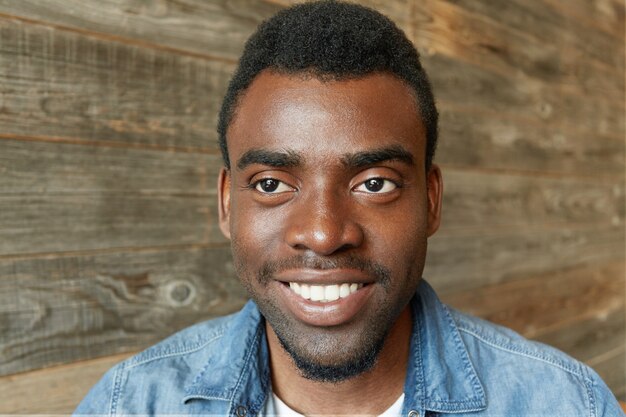 Foto de cabeza de un atractivo joven estudiante de piel oscura con barba en una elegante camisa de jeans sonriendo alegremente mostrando sus perfectos dientes blancos, mirando hacia los lados con una sonrisa inspirada y feliz. Horizontal