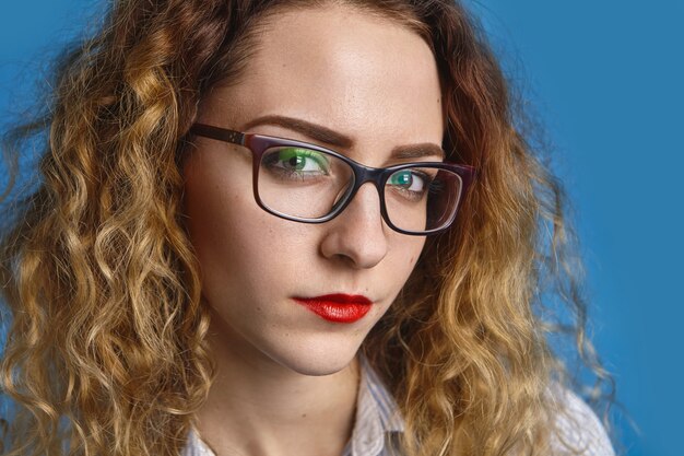 Foto de cabeza de estricta joven empresaria caucásica seria con cabello rizado y labios rojos con expresión facial segura antes de la reunión de negocios, vistiendo camisa formal y anteojos rectangulares