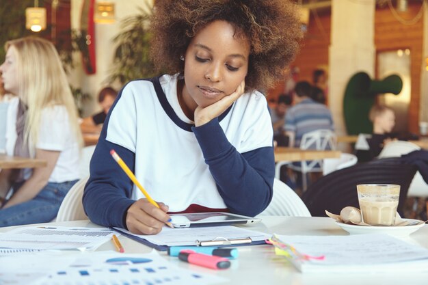 Foto de cabeza de un estudiante africano cansado o aburrido, apoyando la mejilla en la mano mientras trabaja en un proyecto de diploma, usando una conexión a Internet de alta velocidad en el panel táctil, sentada en la cafetería durante el almuerzo