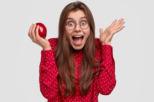 Foto de cabeza de una hermosa joven asombrada extiende las palmas de las manos, se siente asombrado, mira feliz, exclama en voz alta, come una sabrosa manzana jugosa