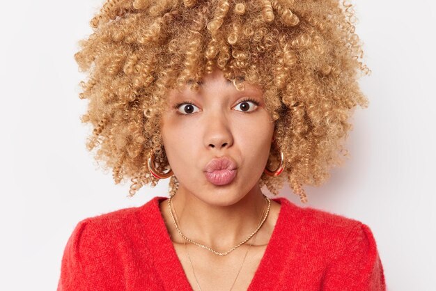 La foto en la cabeza de una hermosa joven romántica con cabello rizado mantiene los labios doblados, sopla el aire, beso a la cámara, mira directamente a la cámara, usa un suéter rojo aislado sobre fondo blanco, te envía mwah