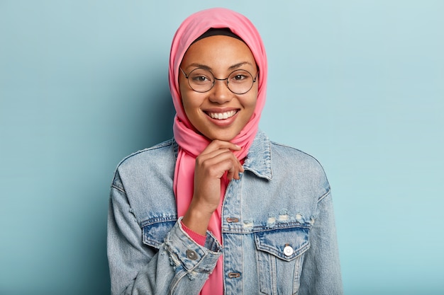 Foto de cabeza de hermosa mujer árabe con una gran sonrisa, sostiene la barbilla, usa gafas redondas, usa ropa tradicional especial