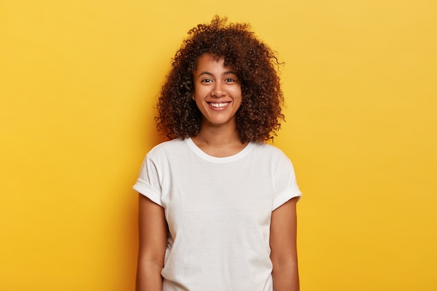 La foto en la cabeza de un hermoso rizado de piel oscura tiene una expresión complacida, se regocija con el éxito, disfruta del tiempo libre, usa una camiseta informal, aislada en la pared amarilla. Personas, emociones positivas, concepto de sentimientos.