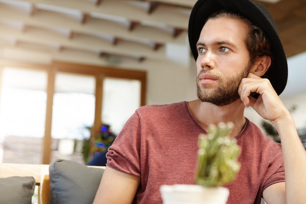 Foto de cabeza de hombre joven de moda con barba con sombrero elegante con expresión pensativa mientras pasa la mañana en un restaurante acogedor, esperando su café y haciendo planes para el día