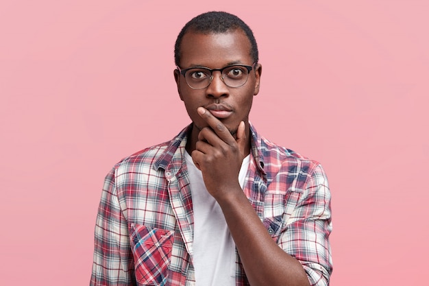 Foto de cabeza de un hombre serio de piel oscura que mira escrupulosamente a la cámara, confiado, usa gafas y camisa a cuadros, aislado sobre rosa