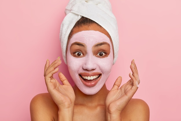 Foto de cabeza de una joven afroamericana optimista que mantiene las manos cerca de la cara, se relaja con una máscara facial, tiene una sonrisa con dientes