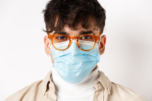 Foto gratuita foto de cabeza de un joven con gafas y una máscara médica estéril que parece alegre y motivado, de pie sobre un fondo blanco. concepto de distanciamiento social y coronavirus