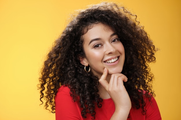 Foto de cabeza de linda mujer romántica femenina tonta y tierna con peinado rizado tocando el labio con el dedo índice haciendo ojos a la cámara y sonriendo como usando habilidades de seducción sobre fondo amarillo.