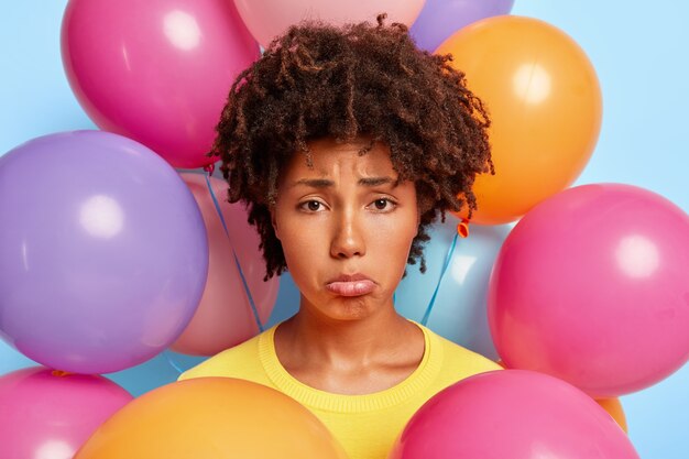 Foto de cabeza de una mujer afro desesperada triste que frunce el labio inferior, está de mal humor durante la fiesta, no tiene amigos, quiere celebrar en gran compañía su cumpleaños, hace una foto cerca de globos de colores. Vacaciones estropeadas