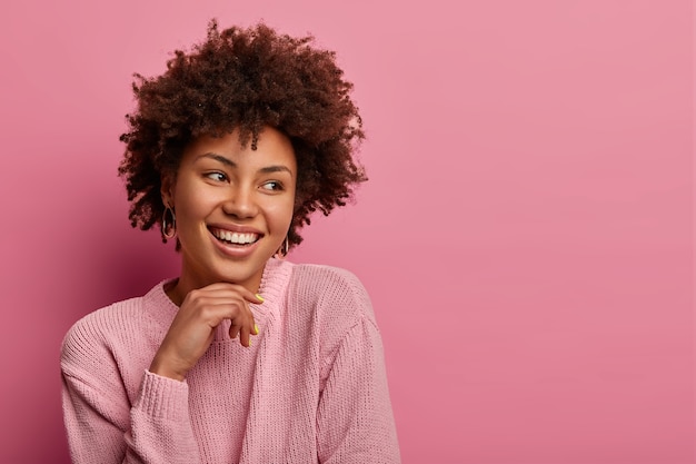 Foto de cabeza de una mujer afro rizada bastante encantadora posa alegre y mira a un lado, mantiene la mano debajo de la barbilla, usa un suéter cálido, se para con una amplia sonrisa, aislado en la pared rosa, espacio en blanco a un lado.