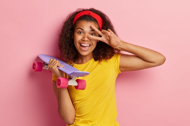 Foto de cabeza de una mujer afroamericana de aspecto agradable que hace un gesto de paz, sonríe positivamente, usa una diadema roja y una camiseta amarilla, sostiene una patineta aislada sobre una pared rosa. Concepto de ocio