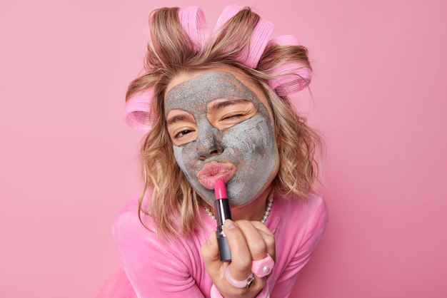 Una foto en la cabeza de una mujer bonita aplica rodillos para el cabello con pintalabios y una máscara de arcilla en la cara se somete a procedimientos de belleza y quiere lucir perfecta en la fiesta o la cita usa ropa festiva aislada sobre fondo rosa