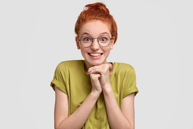 Foto de cabeza de una mujer bonita pelirroja que mira con felicidad, mantiene las manos debajo de la barbilla, anticipa sorpresa preparada por el novio, usa gafas ópticas redondas, camiseta casual, aislado en blanco