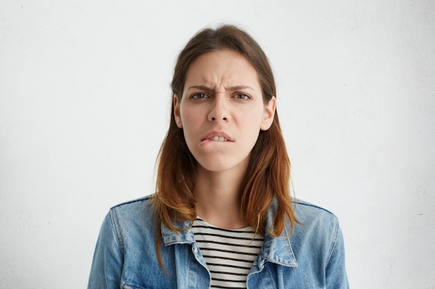 Foto de cabeza de una mujer caucásica joven estresada frustrada vestida con ropa elegante mordiendo el labio inferior y frunciendo el ceño, sintiéndose nerviosa y ansiosa