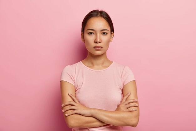 La foto en la cabeza de una mujer coreana seria se ve con expresión tranquila, mantiene los brazos cruzados, tiene una piel fresca y saludable, usa una camiseta rosada, se para en el interior. Hermosa chica asiática tiene mirada segura