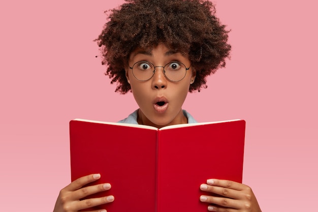 Foto de cabeza de una mujer joven de piel oscura estupefacta que abre la boca del asombro, sostiene un libro de texto rojo, sorprendida con un final inesperado de una historia romántica, modelos de interior sobre una pared rosa. Concepto de Omg