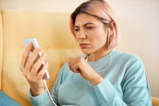 Foto de cabeza de una mujer joven seria con cabello rosado con auriculares, escuchando música en el reproductor multimedia a través del móvil, leyendo un mensaje de texto.