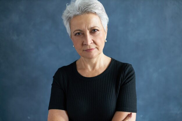 Foto de cabeza de una mujer jubilada de 60 años vestida informalmente con cabello gris que mantiene los brazos cruzados sobre el pecho, mirando a la cámara con una mirada sospechosa escrutadora, entrecerrando los ojos