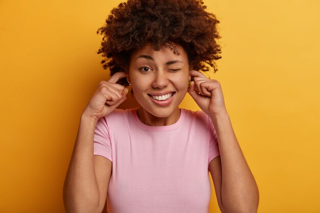 La foto en la cabeza de una mujer muy feliz muestra los dientes, mantiene los dedos índices en los orificios de las orejas, usa ropa informal, posa sobre una pared amarilla, no puede concentrarse en un lugar tan ruidoso, guiña los ojos, evita el sonido fuerte