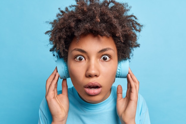 La foto en la cabeza de una mujer de pelo rizado impresionada y aturdida tiene los ojos abiertos mantiene el aliento escucha música en auriculares inalámbricos sorprendido al escuchar un sonido muy fuerte inesperadamente aislado en una pared azul