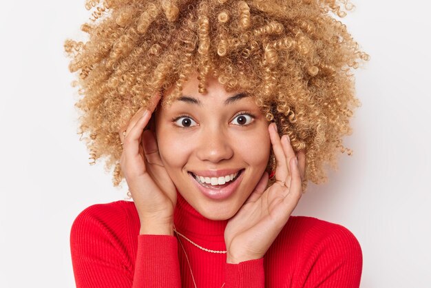 Foto de cabeza de mujer de pelo rizado toca la cara sonríe ampliamente se siente feliz viste un cuello de tortuga rojo casual rojo aislado sobre fondo blanco se siente alegre reacciona en un presente impresionante. Concepto de emociones.