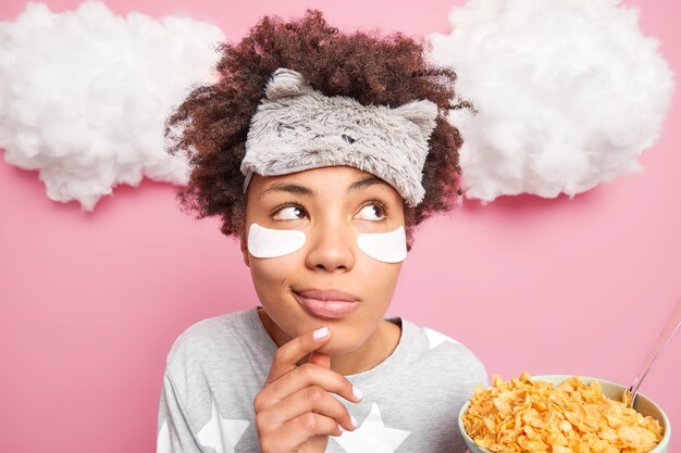 Foto de cabeza de una mujer rizada soñadora reflexiva mantiene la mano en la barbilla se ve pensativa a un lado vestida en pijama tiene un desayuno saludable sostiene un tazón de copos de maíz hace planes para el día de trabajo