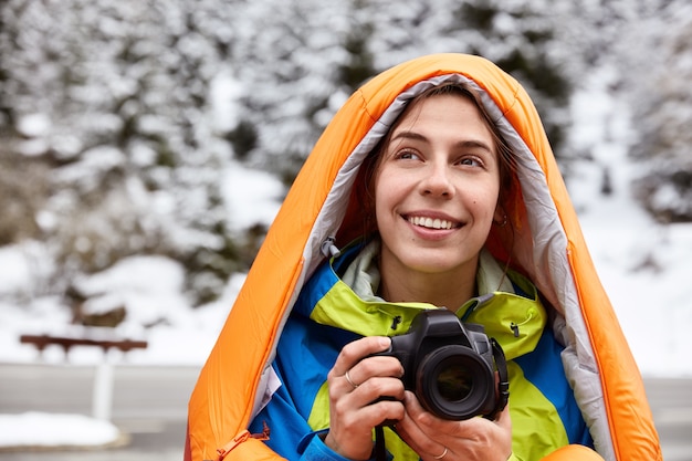 Foto de cabeza de una viajera alegre que se recrea en montañas nevadas, hace fotos de vistas panorámicas, posa contra el espacio invernal, sonríe suavemente