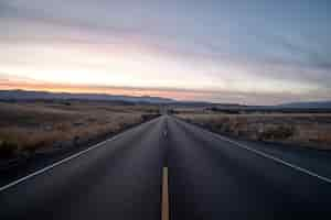 Foto gratuita foto de una carretera rodeada de campos de hierba seca bajo un cielo durante la puesta de sol