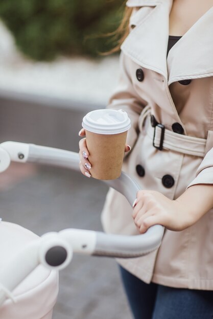 Foto de cerca, mujer sosteniendo una taza de café desechable y quedándose en el cochecito de bebé.