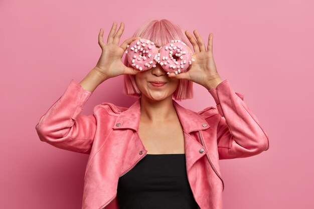 La foto de una chica asiática de pelo rosa de moda cubre los ojos con deliciosas donas, disfruta de una sabrosa repostería aromática, come donas glaseadas